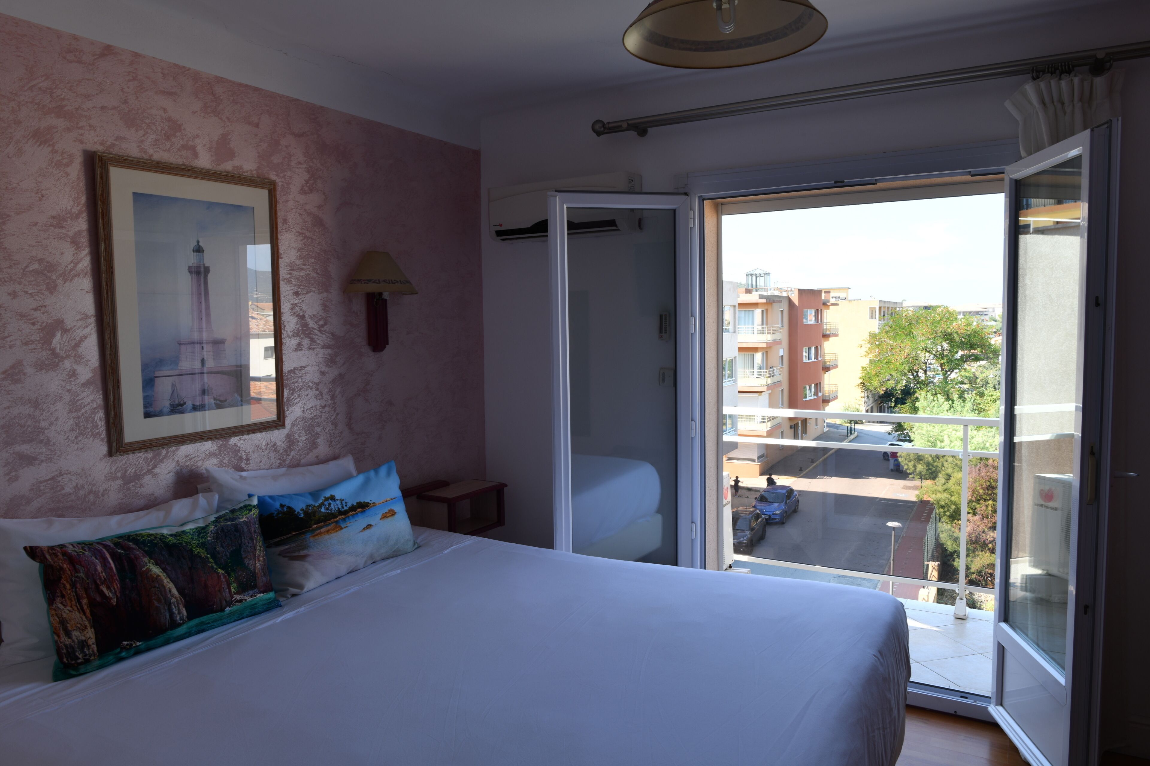 A bedroom with a bed, pillows, a painting, a nightstand, and a sliding glass door leading to a balcony with a view of buildings and a street.