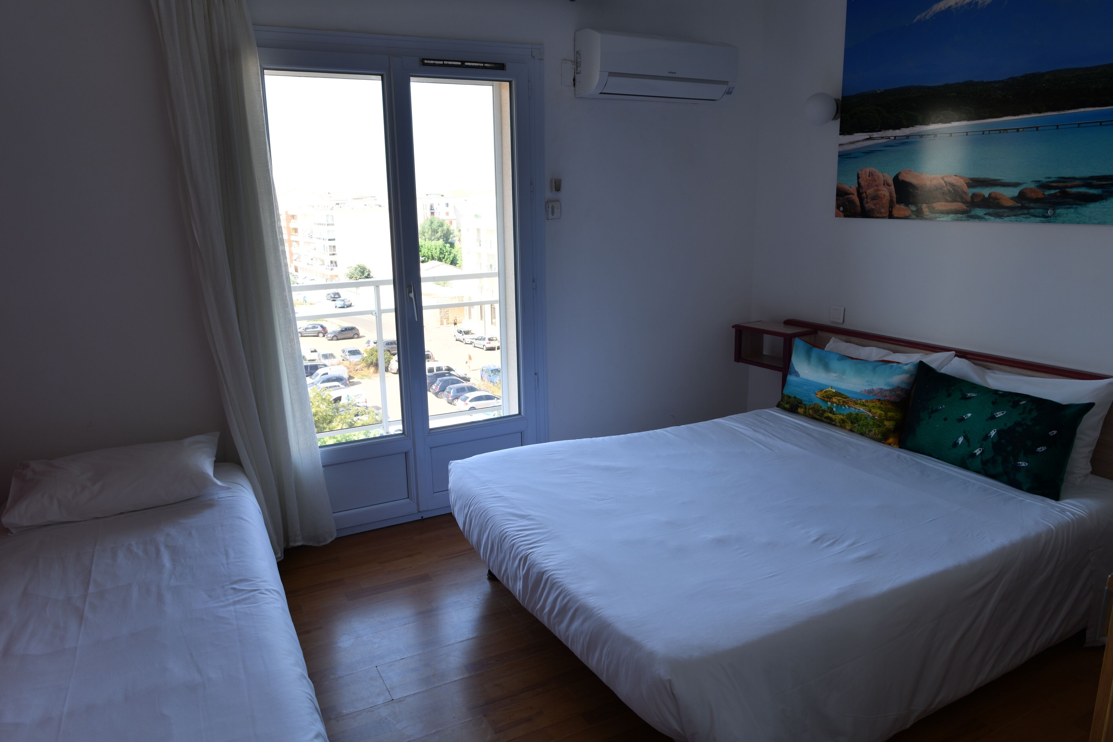 Een slaapkamer met een groot bed, een kleiner bed, een raam met gordijnen en een schilderij aan de muur.