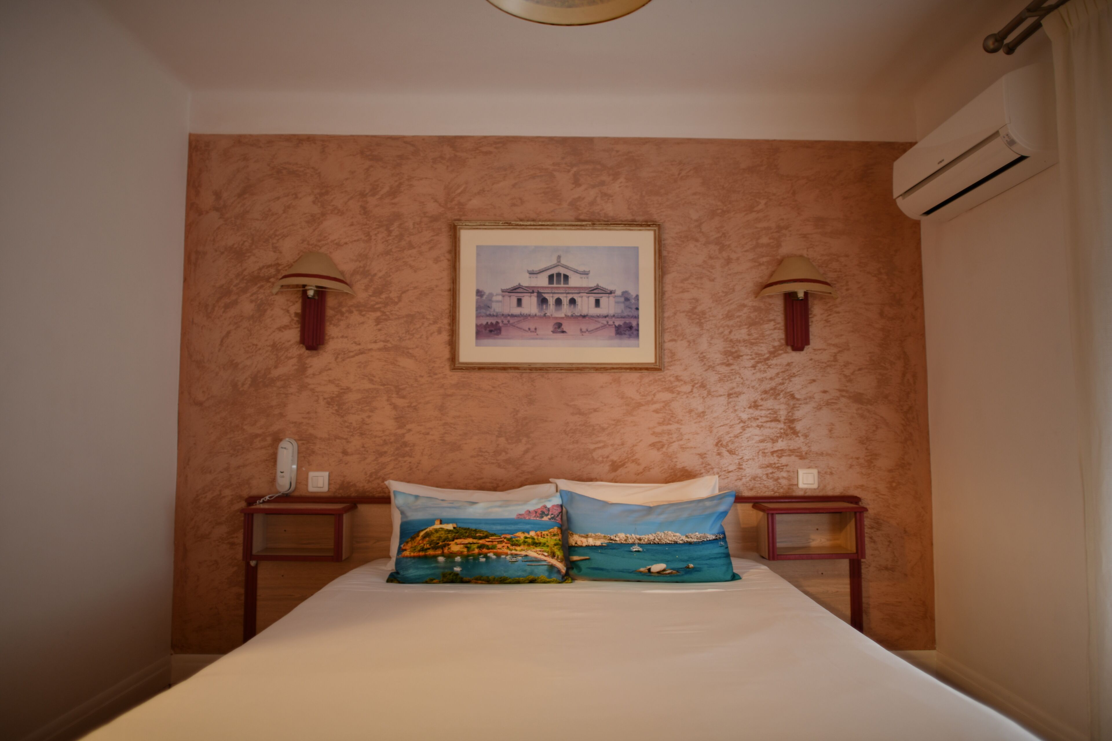 A bedroom with a bed, two pillows with landscape prints, a framed picture on the wall, two bedside lamps, and an air conditioning unit.