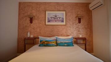 A bedroom with a bed, two pillows with landscape prints, a framed picture on the wall, two bedside lamps, and an air conditioning unit.