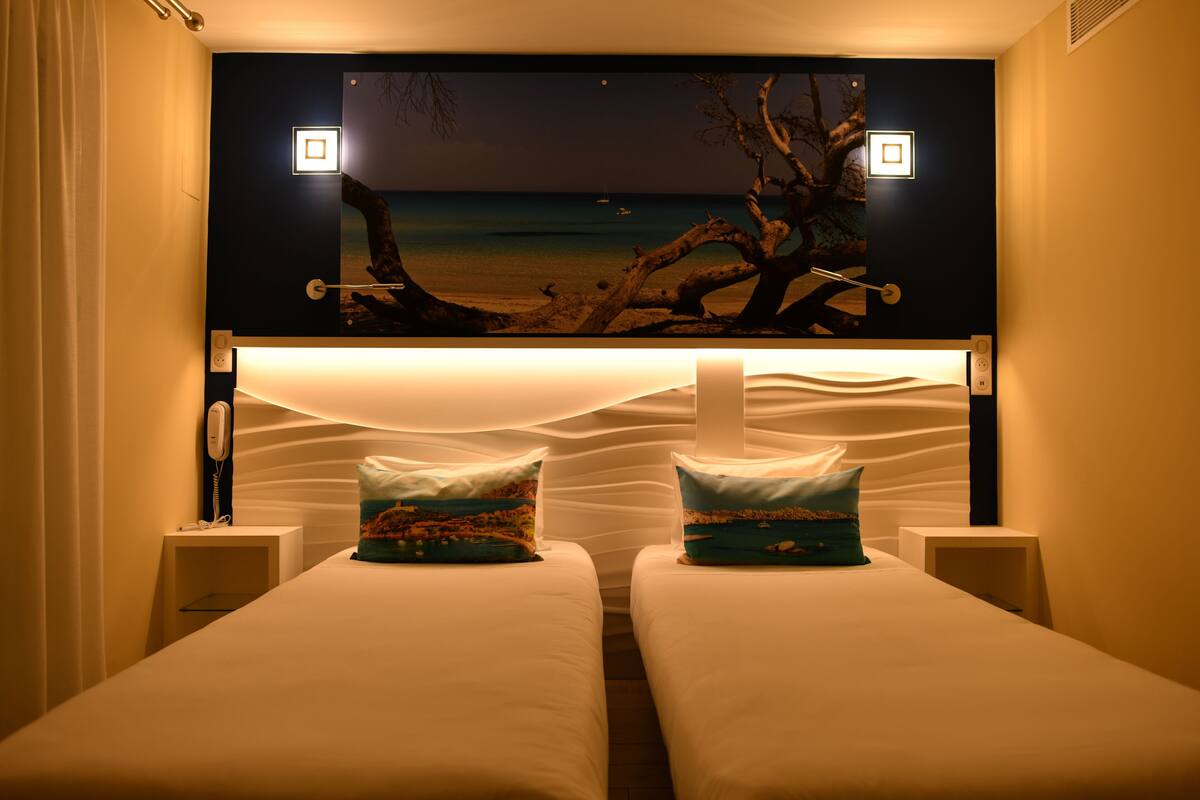 A hotel room with two single beds, a nightstand with a phone, and a large wall-mounted artwork depicting a beach scene.