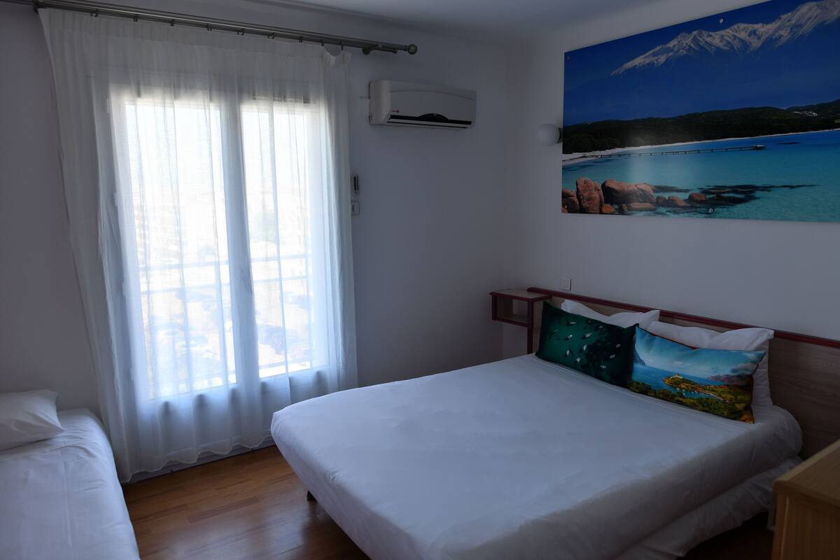 A bedroom with a bed, a window with sheer curtains, an air conditioning unit, and a painting of a coastal landscape.