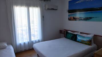 Une chambre à coucher avec un lit, une fenêtre ornée de rideaux légers, un appareil de climatisation et un tableau représentant un paysage côtier.