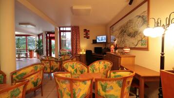 A dining area with wooden chairs and a table, a television on a desk, and a large painting on the wall.