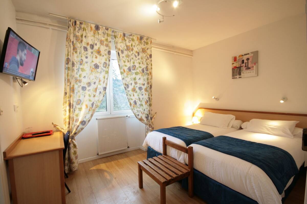 Habitación de hotel con cama, escritorio, silla, televisión y ventana con cortinas florales.