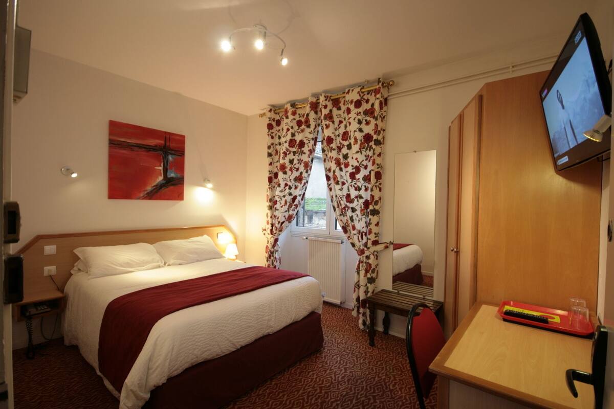 Habitación de hotel con cama, escritorio con teclado, televisor y ventana con cortinas florales.