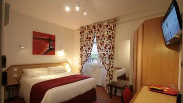 A hotel room with a bed, a desk with a keyboard, a TV, and a window with floral curtains.