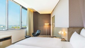 Hotelkamer met een groot bed, een bureau, een stoel, een vloerlamp en uitzicht op de stad door grote ramen.