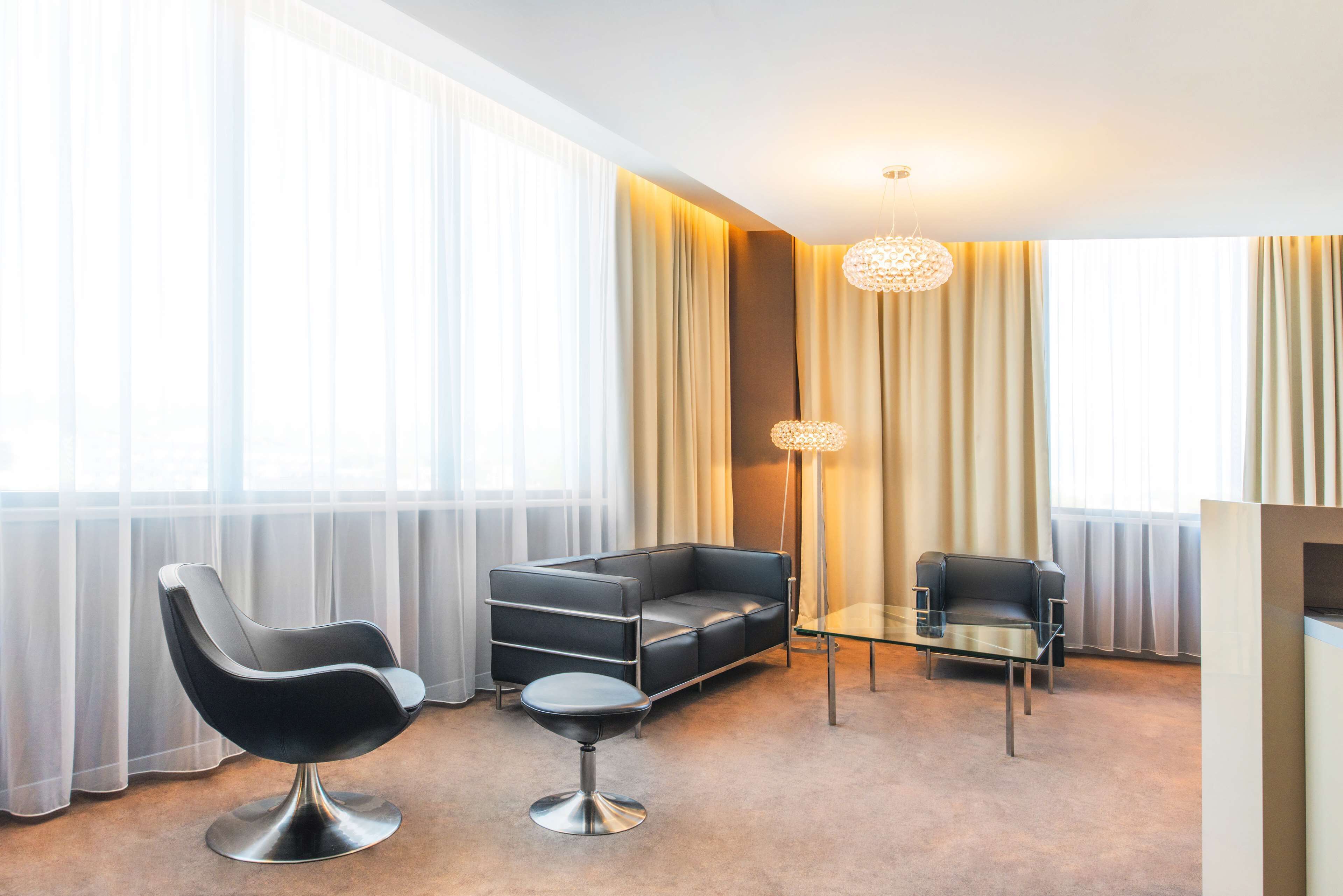 A modern hotel room with a black leather sofa, a black leather chair, a glass coffee table, and a floor lamp.
