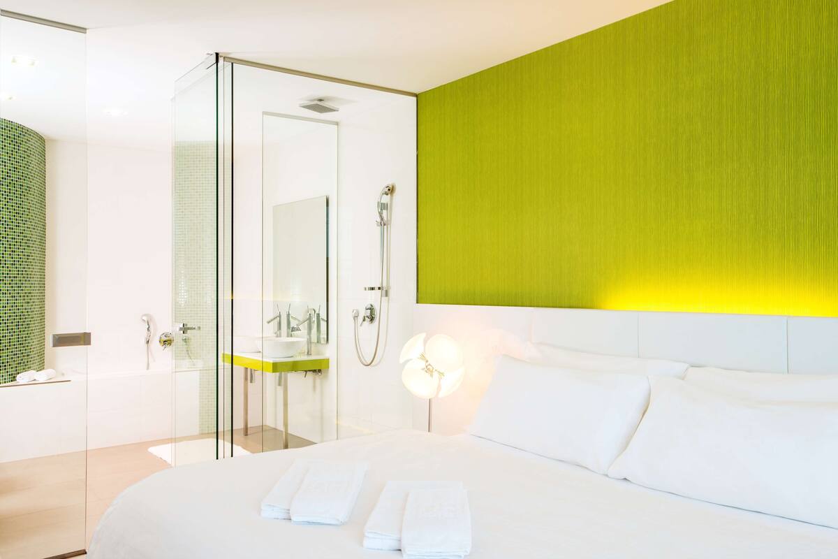 A modern hotel room with a large bed, a glass shower enclosure, and a green accent wall.