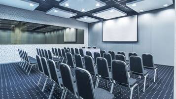Un salón de conferencias con filas de sillas, una pantalla de proyector y luces en el techo.