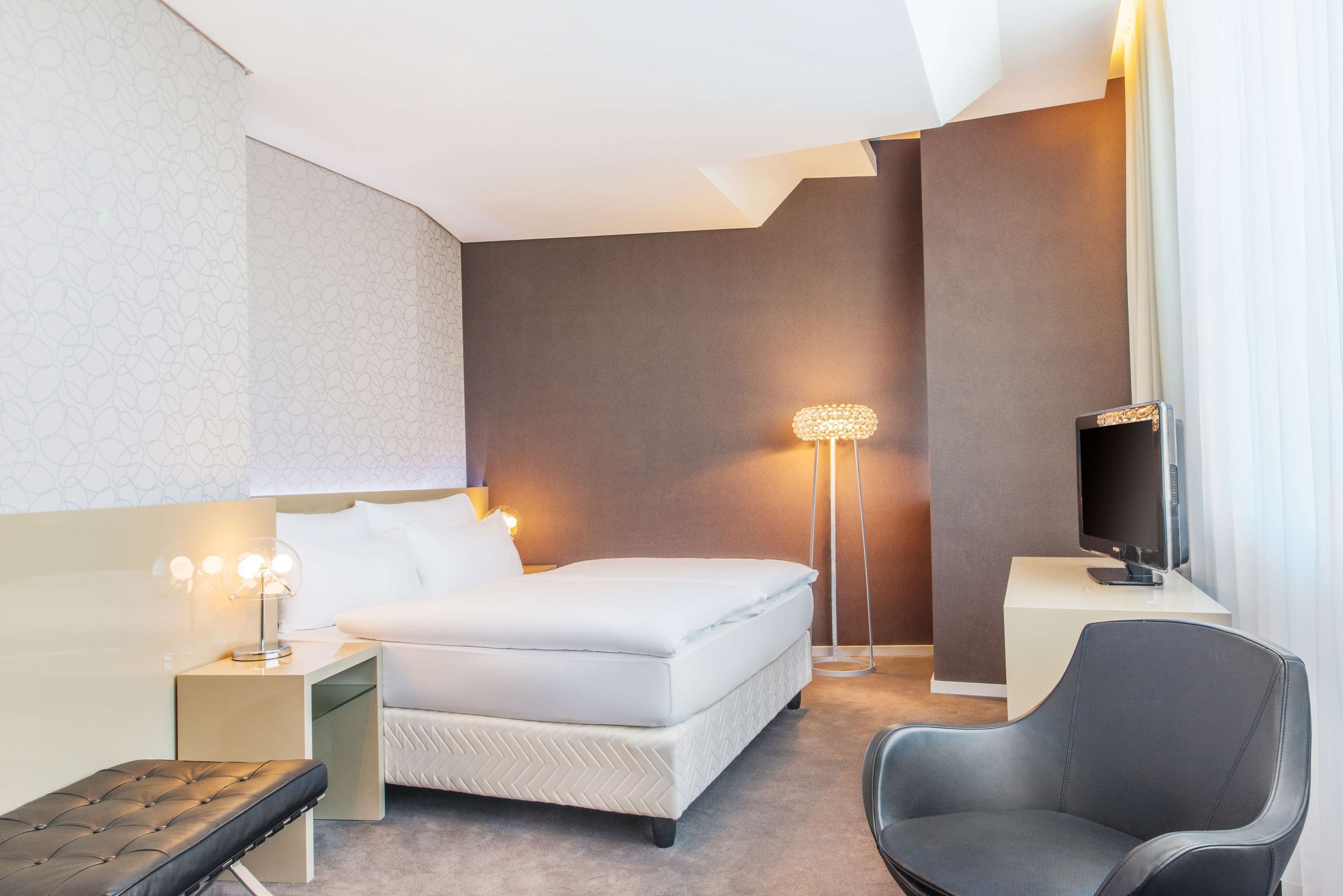 Una habitación de hotel moderna con cama, lámpara de noche, sillón y televisor de pantalla plana.
