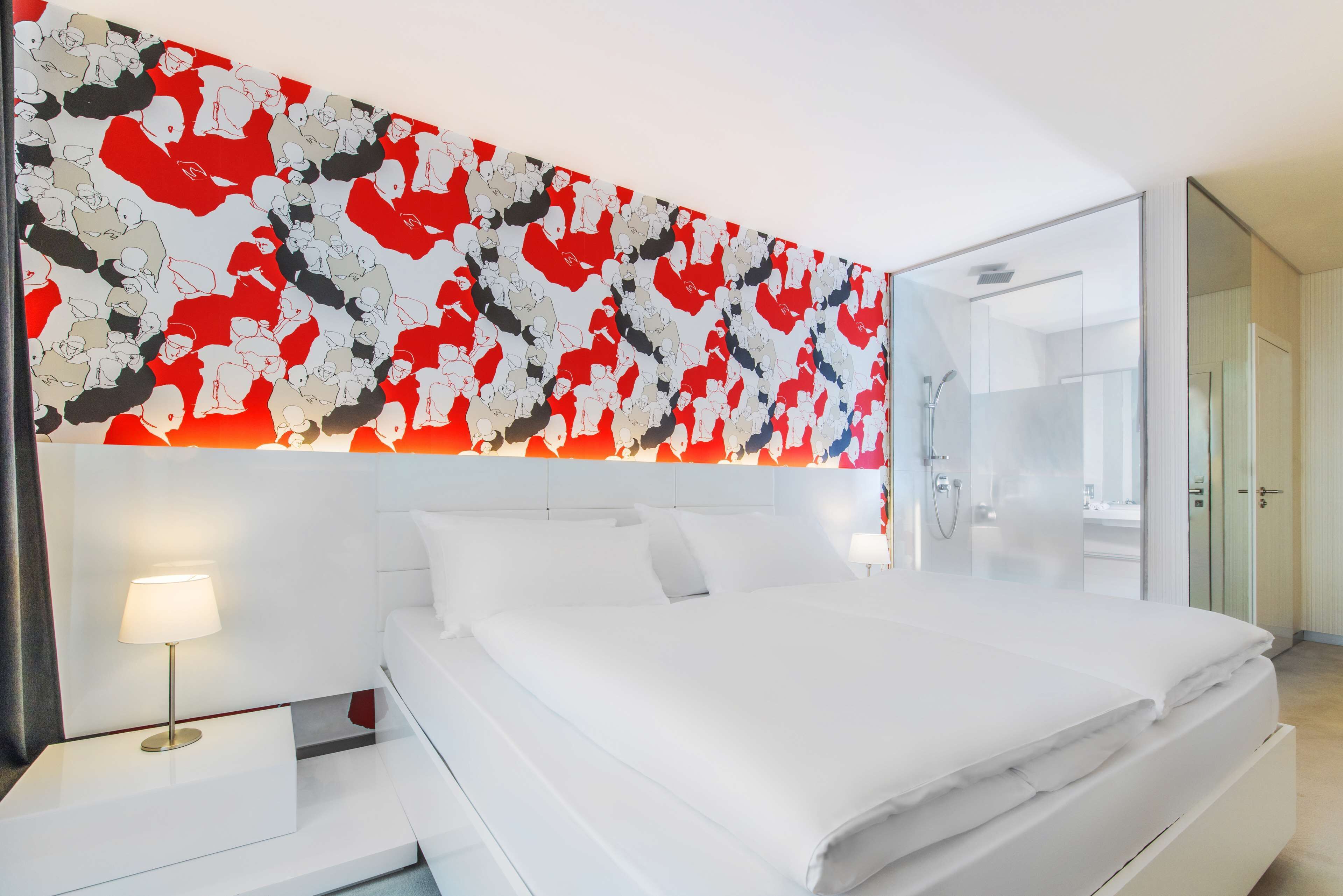 Une chambre d’hôtel moderne avec un grand lit, du linge de lit blanc, une table de chevet avec une lampe et une œuvre d’art abstraite et colorée au-dessus du lit.