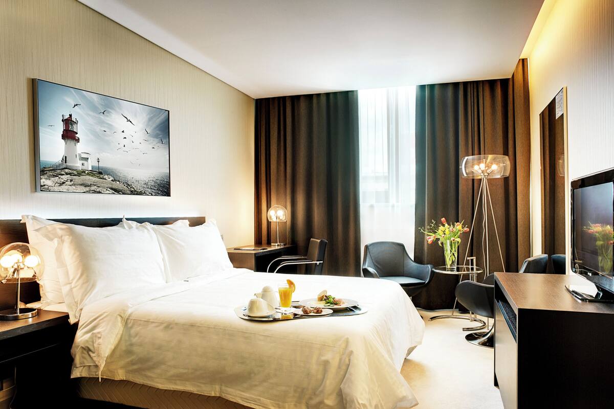 Habitación de hotel con cama, mesita de noche, escritorio, silla, televisor y un cuadro de un faro.