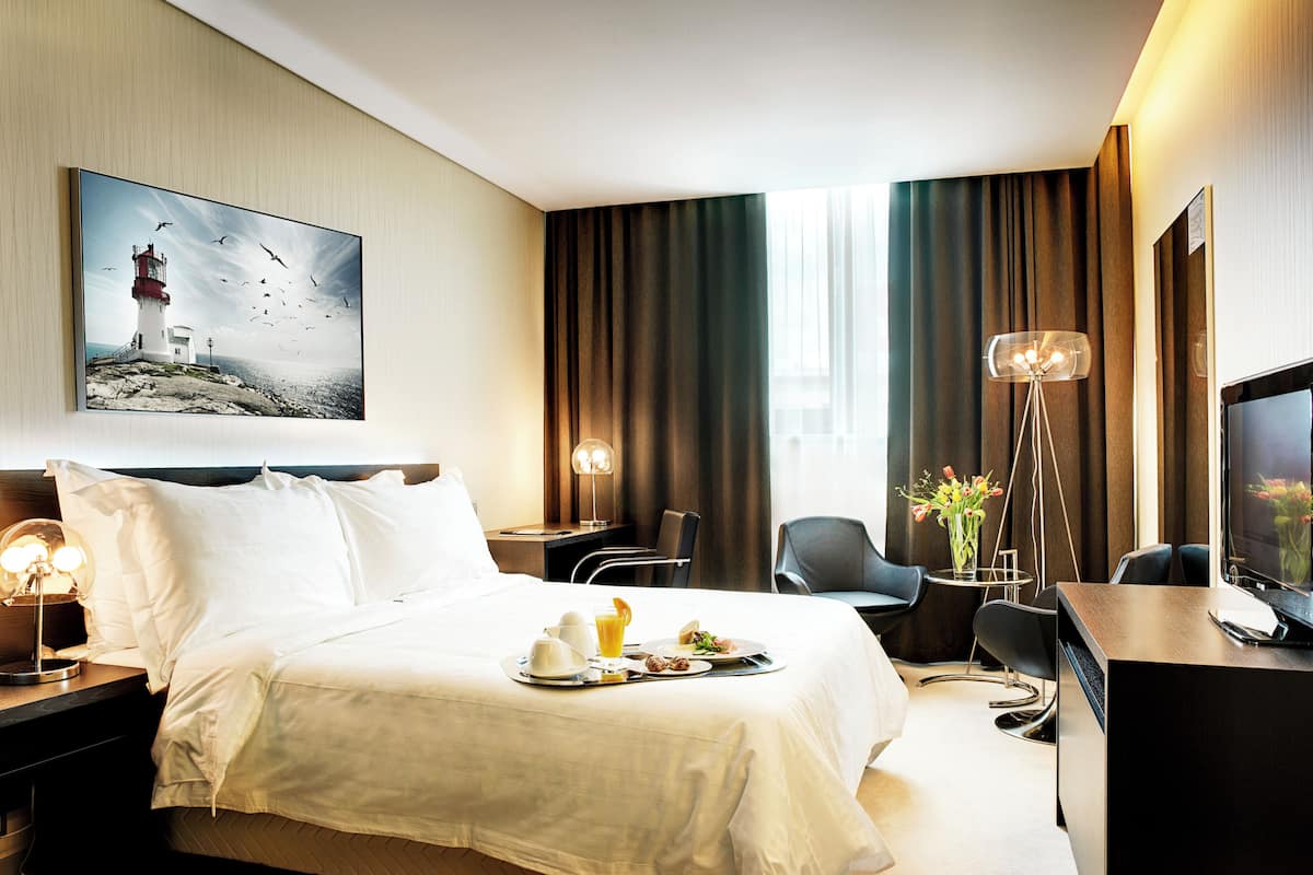 A hotel room with a bed, bedside table, desk, chair, TV, and a painting of a lighthouse.