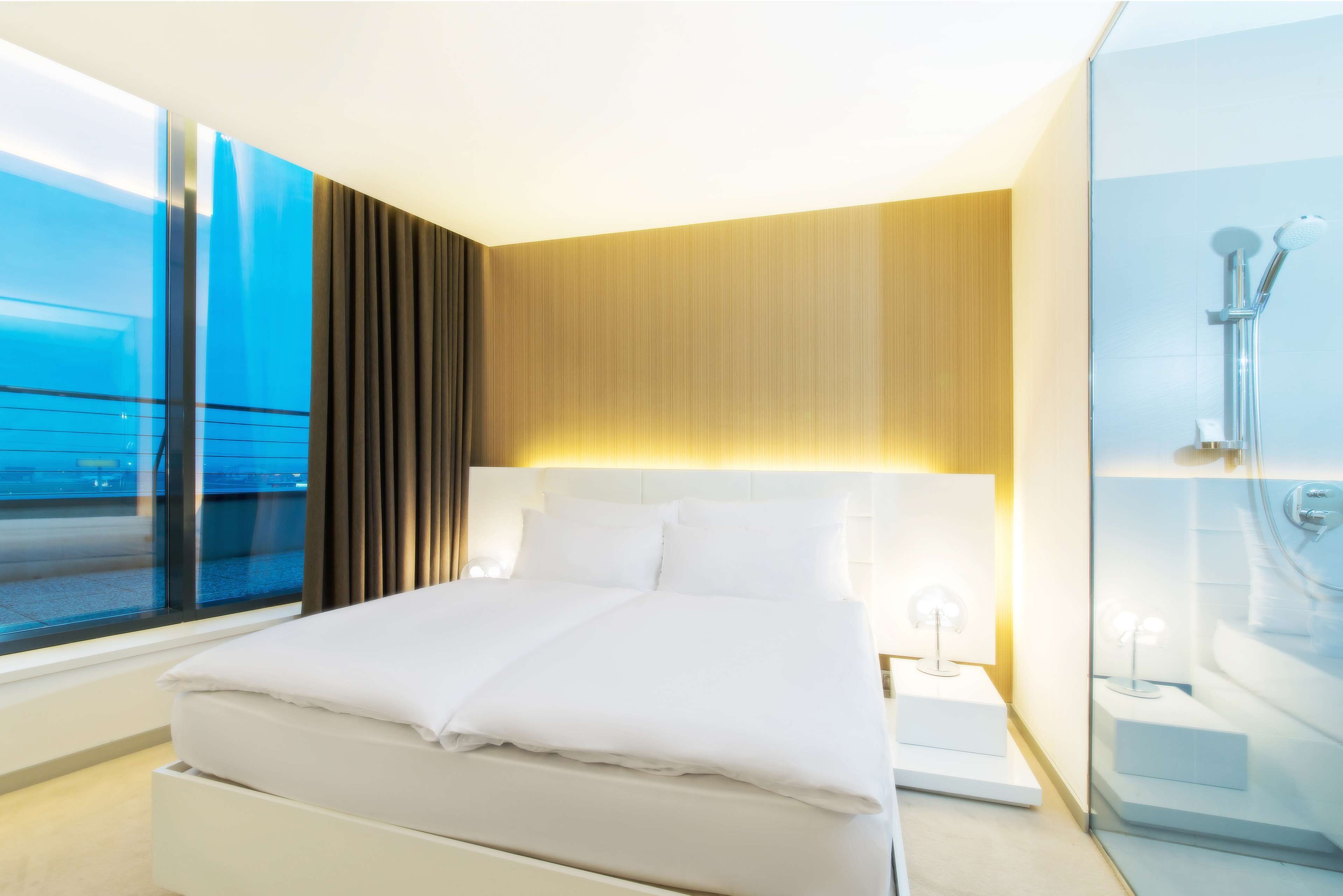 Een moderne hotelkamer met een groot bed, een nachtkastje, een glazen douchecabine en uitzicht op zee door een groot raam.