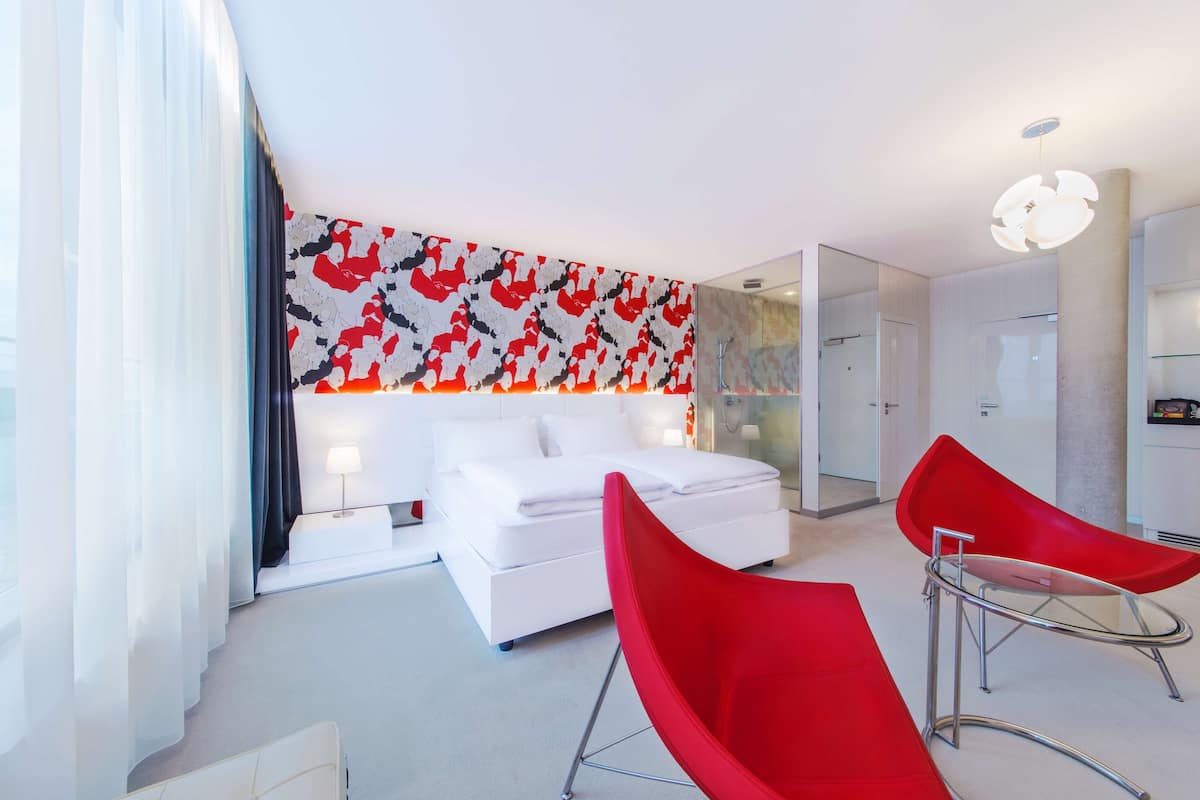 A modern hotel room with a white bed, red chairs, and a large abstract wall art piece.