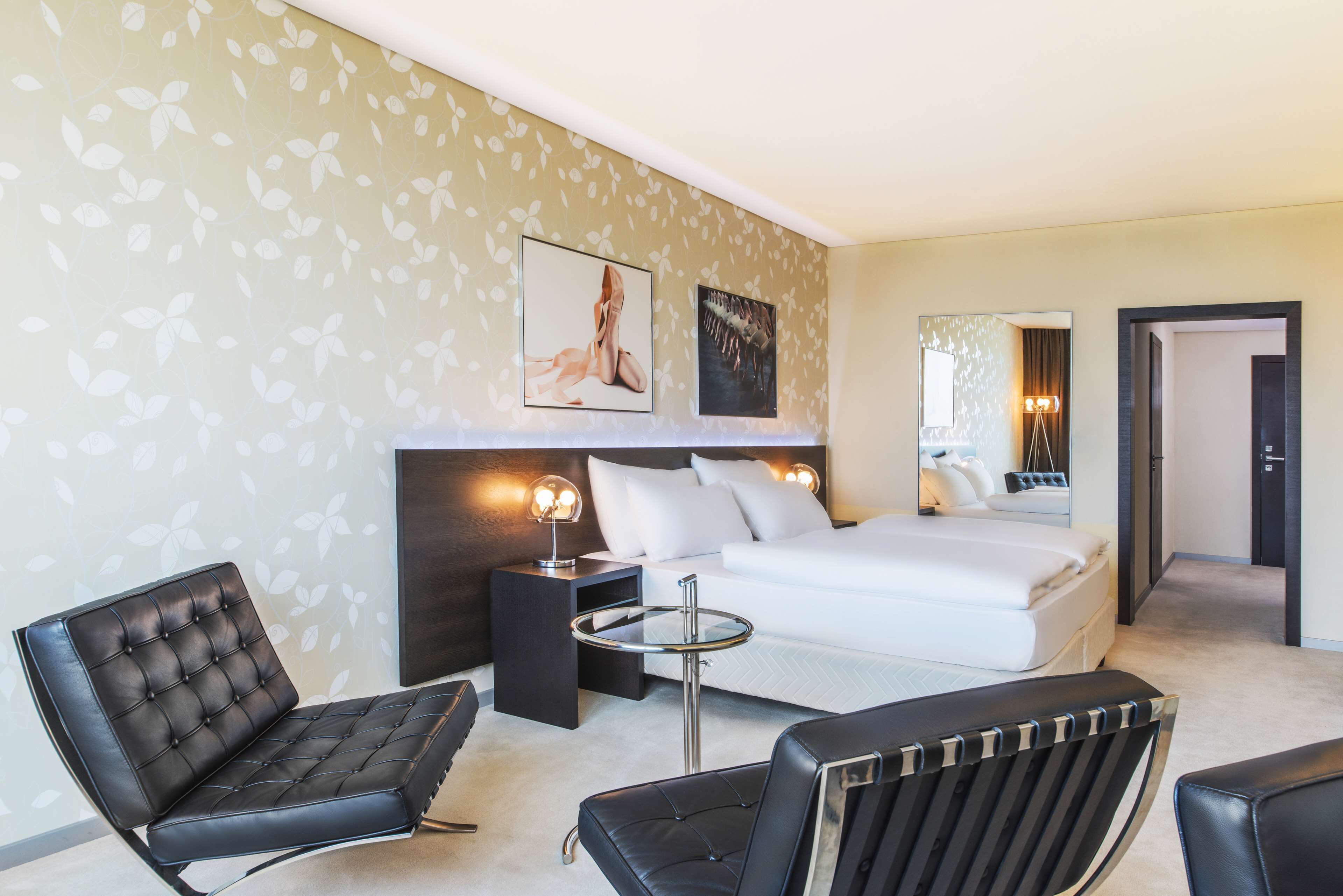 Una habitación de hotel con una cama, dos sillones de cuero negro, una mesita redonda y una mesita de noche con una lámpara.