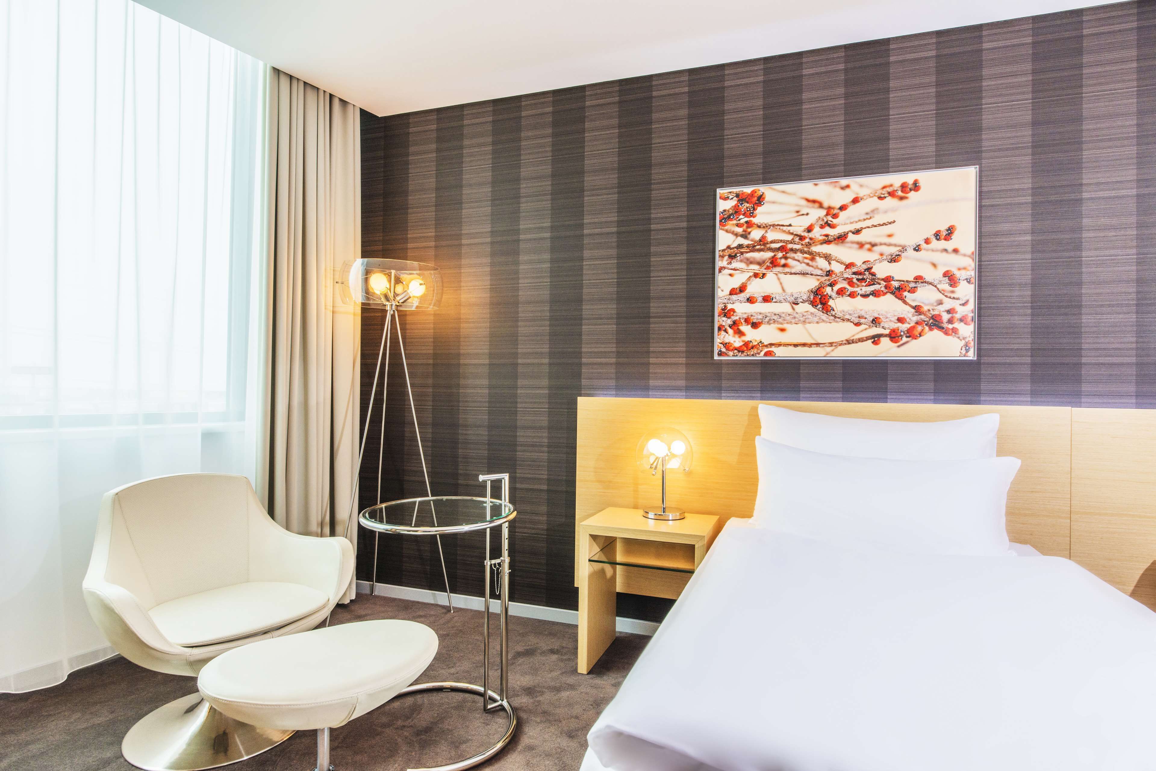 Een moderne hotelkamer met een bed, een stoel, een nachtkastje met een lamp, een vloerlamp en een groot raam met gordijnen.