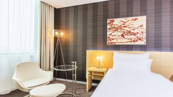 Een moderne hotelkamer met een bed, een stoel, een nachtkastje met een lamp, een vloerlamp en een groot raam met gordijnen.