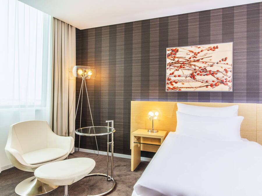 Een moderne hotelkamer met een bed, een stoel, een nachtkastje met een lamp, een vloerlamp en een groot raam met gordijnen.