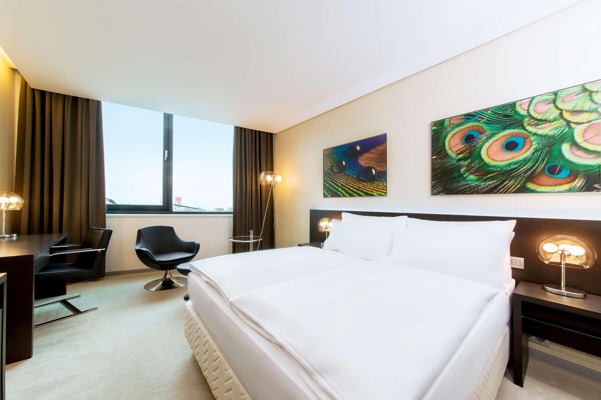 Habitación de hotel con cama, escritorio, silla, ventana y dos cuadros colgados en la pared.
