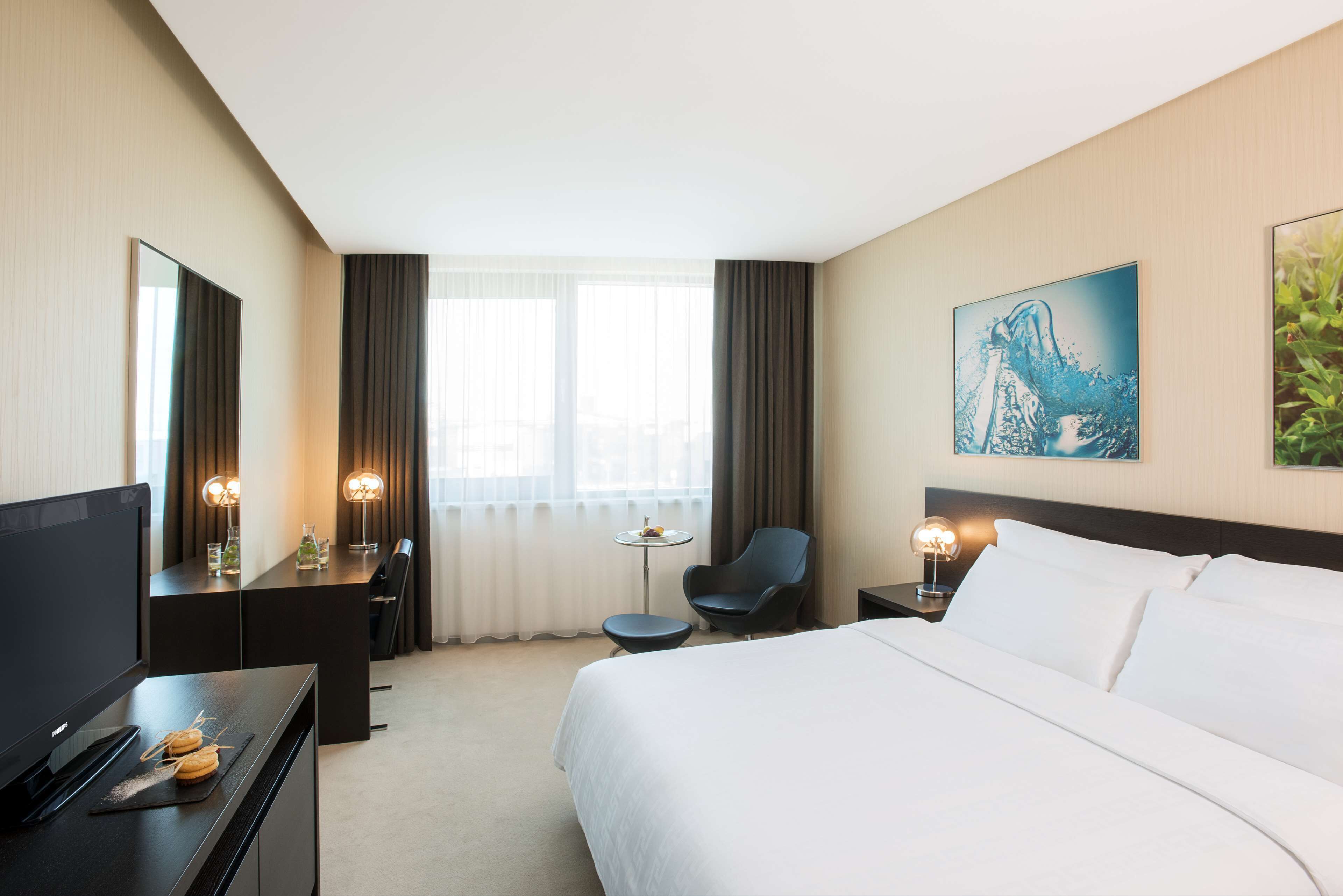 Een hotelkamer met een groot bed, een bureau met een televisie, een stoel, een klein tafeltje met een lamp en een schilderij aan de muur.