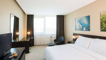 Een hotelkamer met een groot bed, een bureau met een televisie, een stoel, een klein tafeltje met een lamp en een schilderij aan de muur.