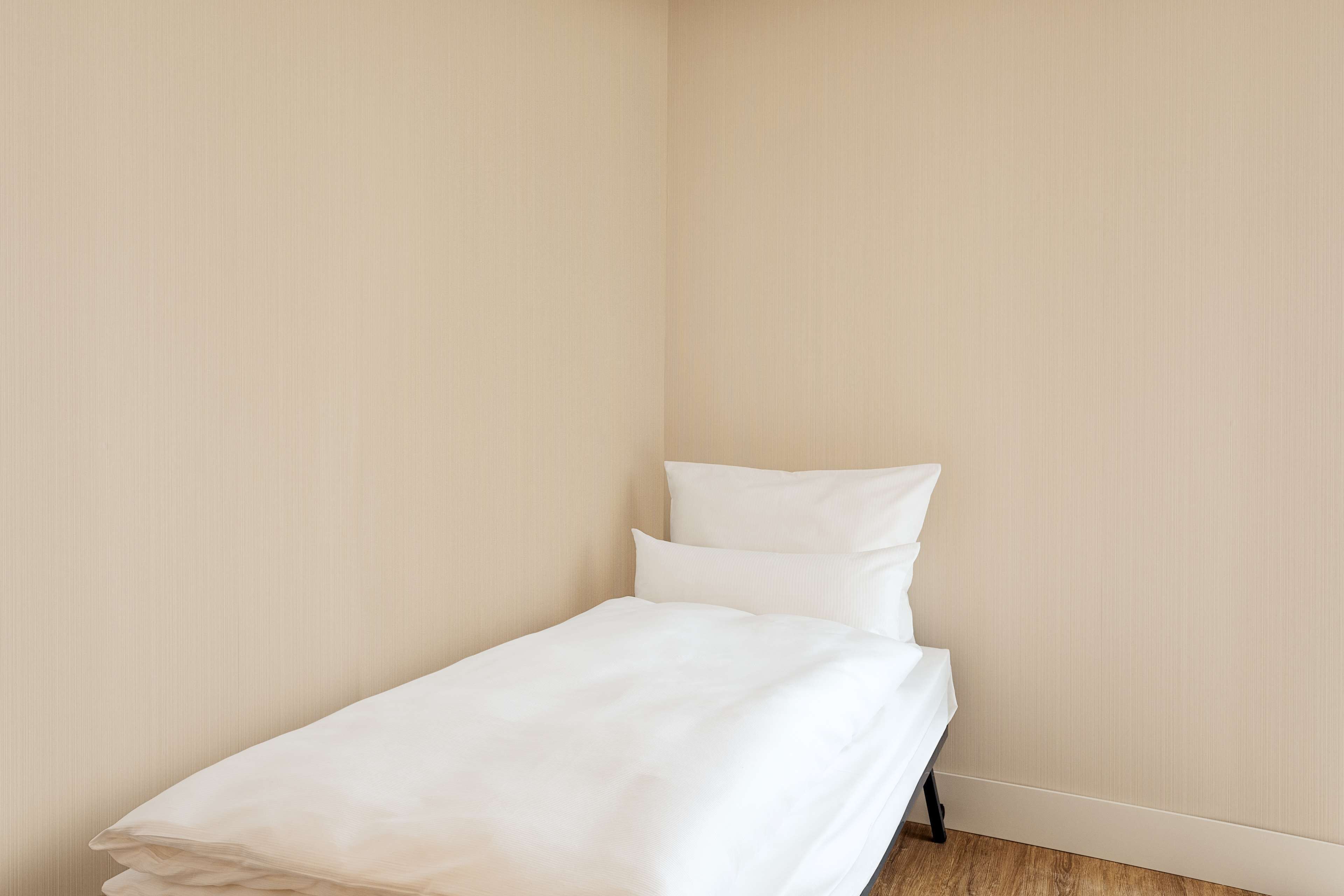 Un petit lit bien fait, avec une literie blanche, dans une chambre d’angle aux murs sobres et au plancher en bois.