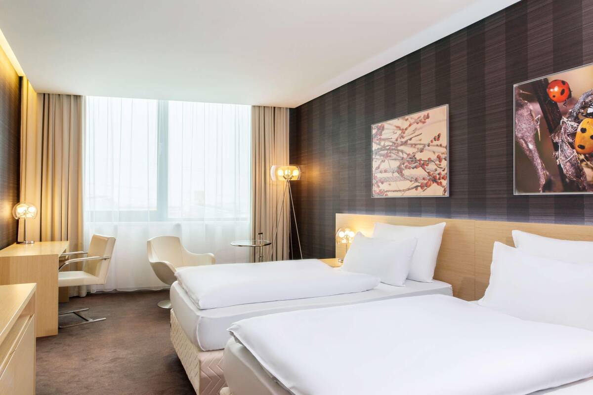 A hotel room with two beds, a desk, a chair, a lamp, and artwork on the wall.