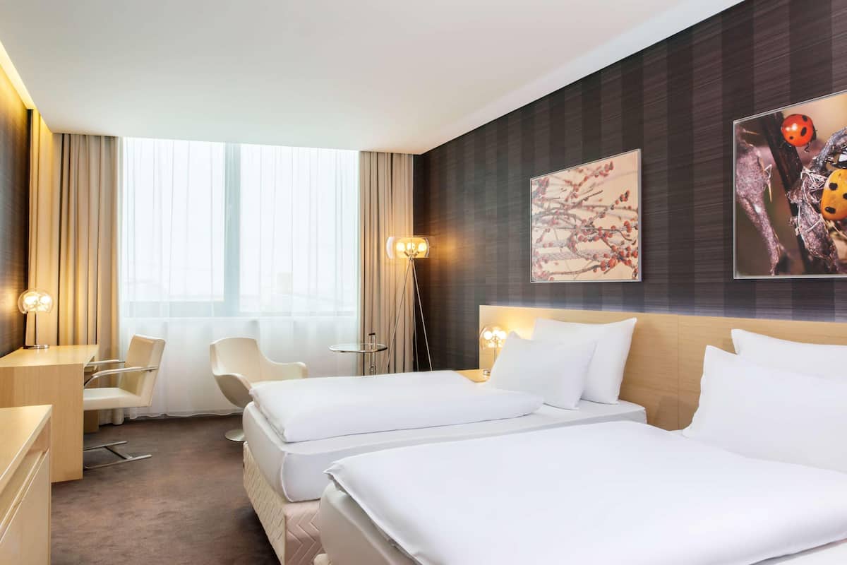 A hotel room with two beds, a desk, a chair, a lamp, and artwork on the wall.