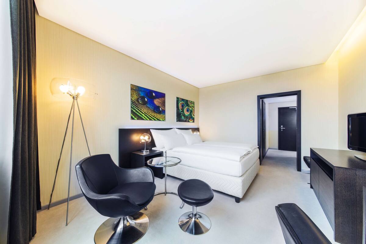 A hotel room with a bed, a black chair, a small table, a desk, and a TV.
