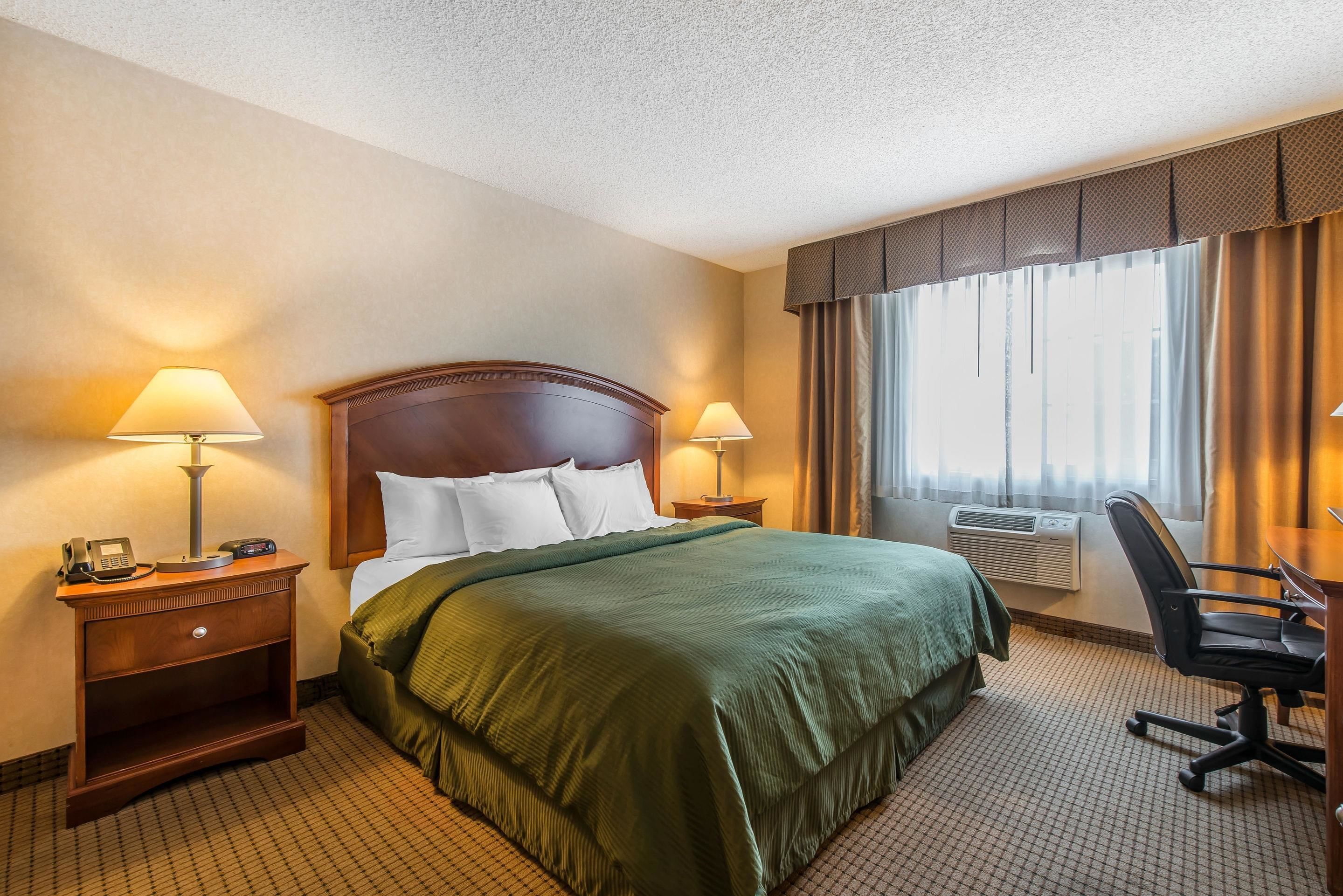 A hotel room with a large bed, two bedside tables with lamps, a desk with a chair, and a window with curtains.