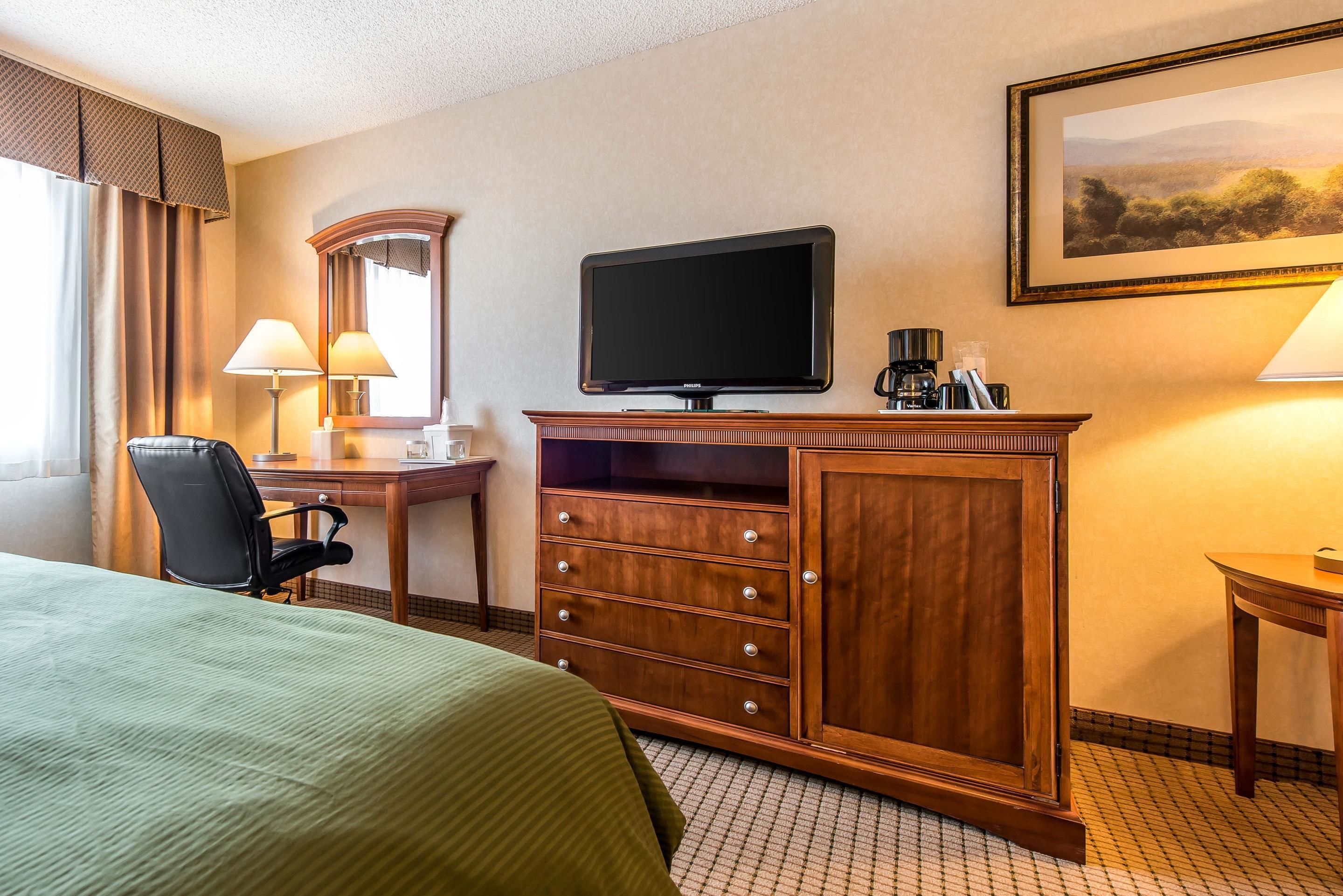 A hotel room with a bed, a desk with a chair, a television, a dresser, and a framed picture on the wall.