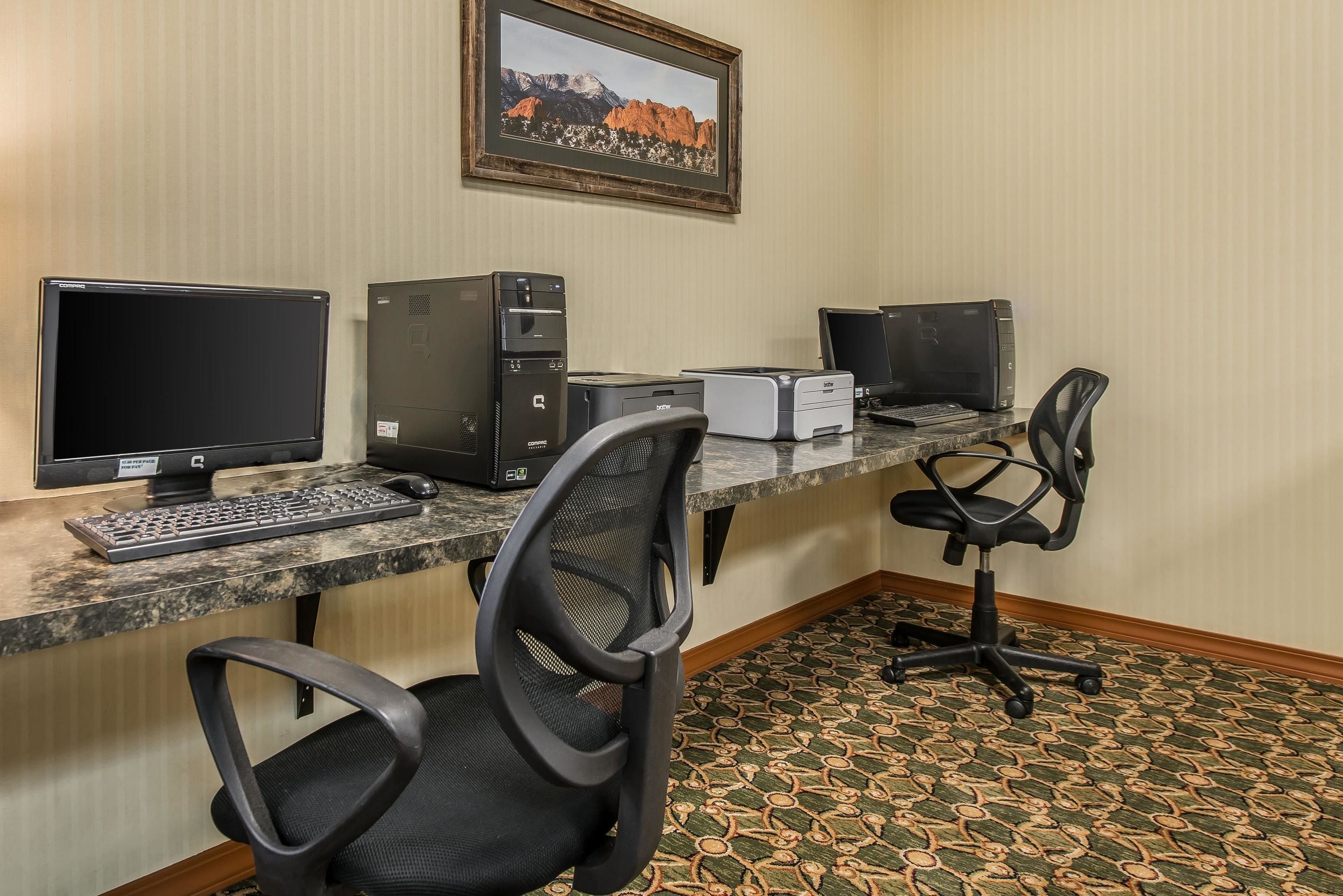 A hotel workspace with two desktop computers, a printer, a keyboard, and two office chairs.