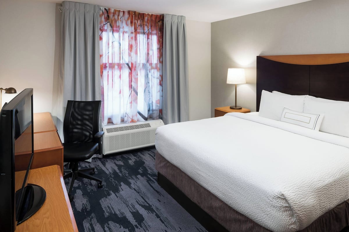 Ein Hotelzimmer mit einem Bett, einem Schreibtisch mit Computer, einem Stuhl, einer Lampe und einem Fenster mit Vorhängen.