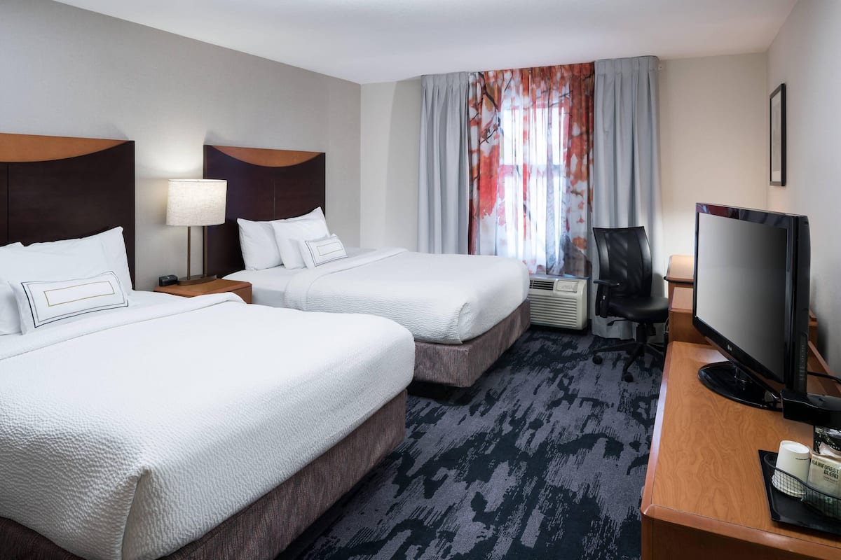 Ein Hotelzimmer mit zwei Betten, einem Schreibtisch mit Computer, einem Fernseher und einem Fenster mit Vorhängen.
