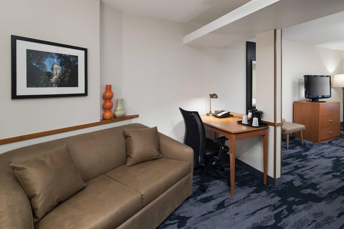 Ein Hotelzimmer mit braunem Sofa, Schreibtisch mit Stuhl, Fernseher und einem gerahmten Bild an der Wand.
