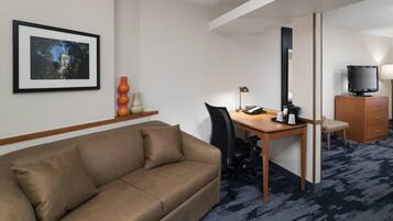 A hotel room with a brown sofa, a desk with a chair, a television, and a framed picture on the wall.