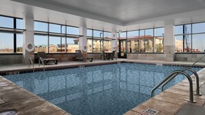 Una amplia piscina interior con agua cristalina y azul, rodeada de sillones y mesas, y grandes ventanales que ofrecen vistas a edificios exteriores.