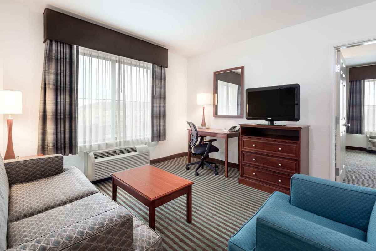 A hotel room with a sofa, a desk with a chair, a television, a dresser, and a large window with curtains.