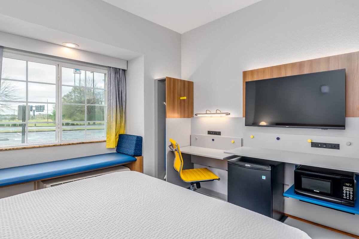 A modern hotel room with a large window, a flat-screen TV, a microwave, and a small kitchenette.