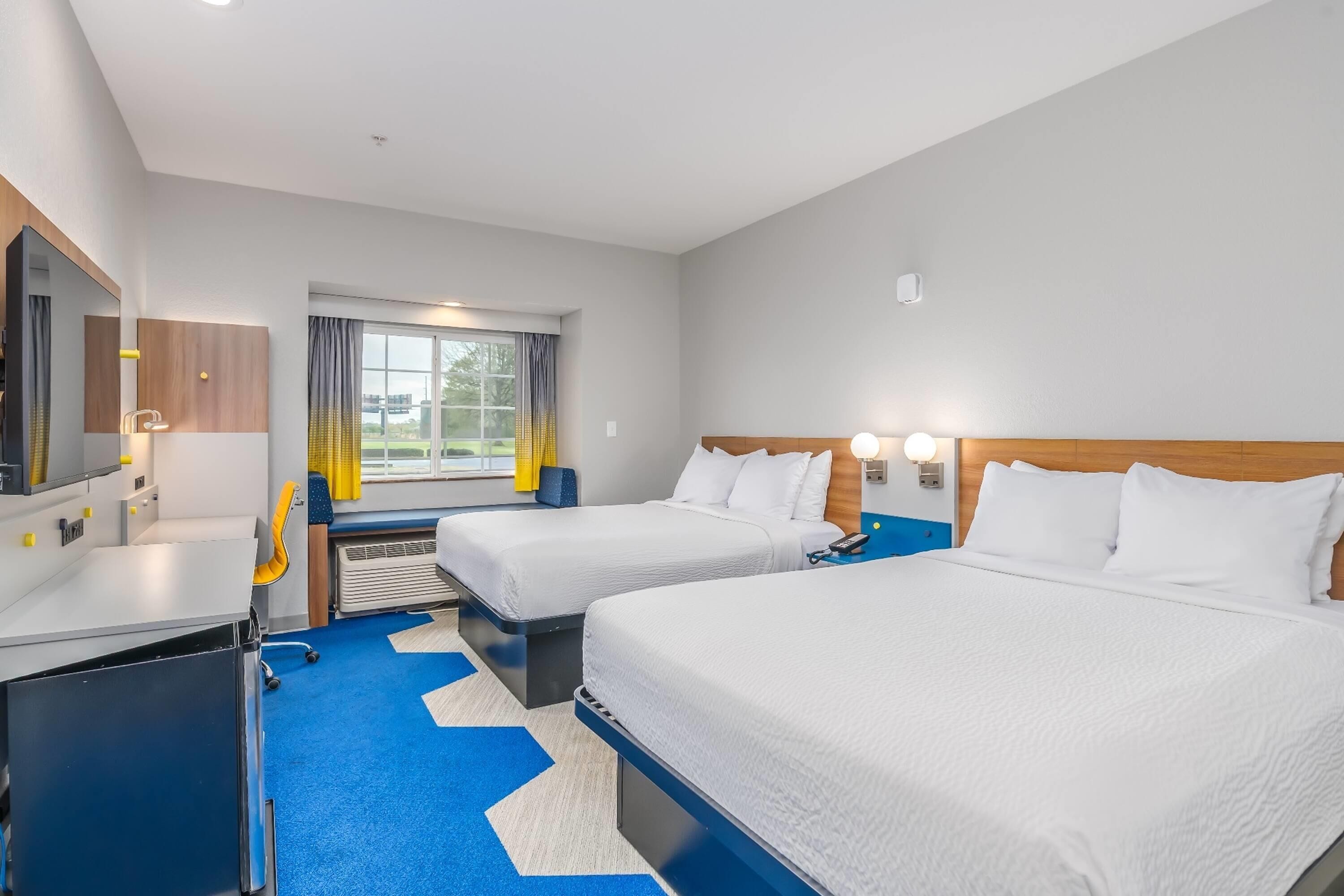 A hotel room with two beds, a desk, a chair, a window with curtains, and a view of the outdoors.