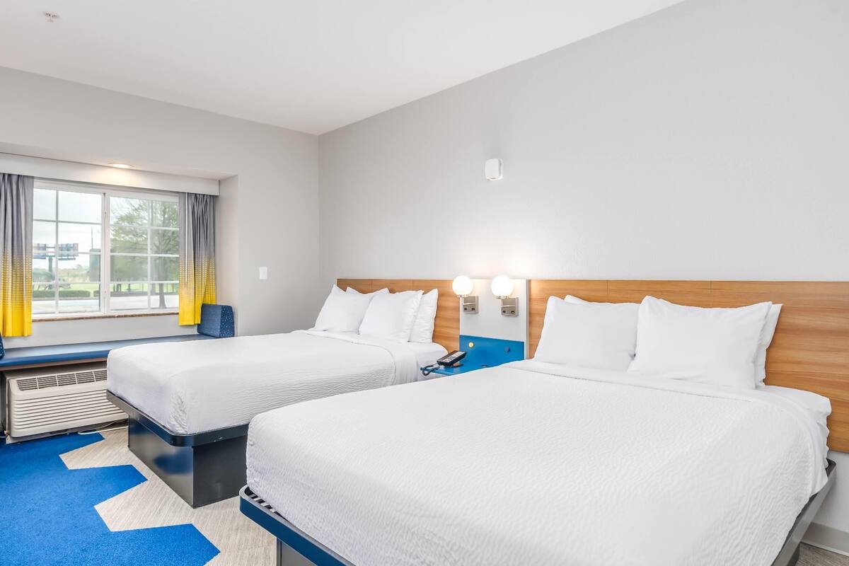A hotel room with two beds, a window with curtains, a heater, and a wall-mounted phone.