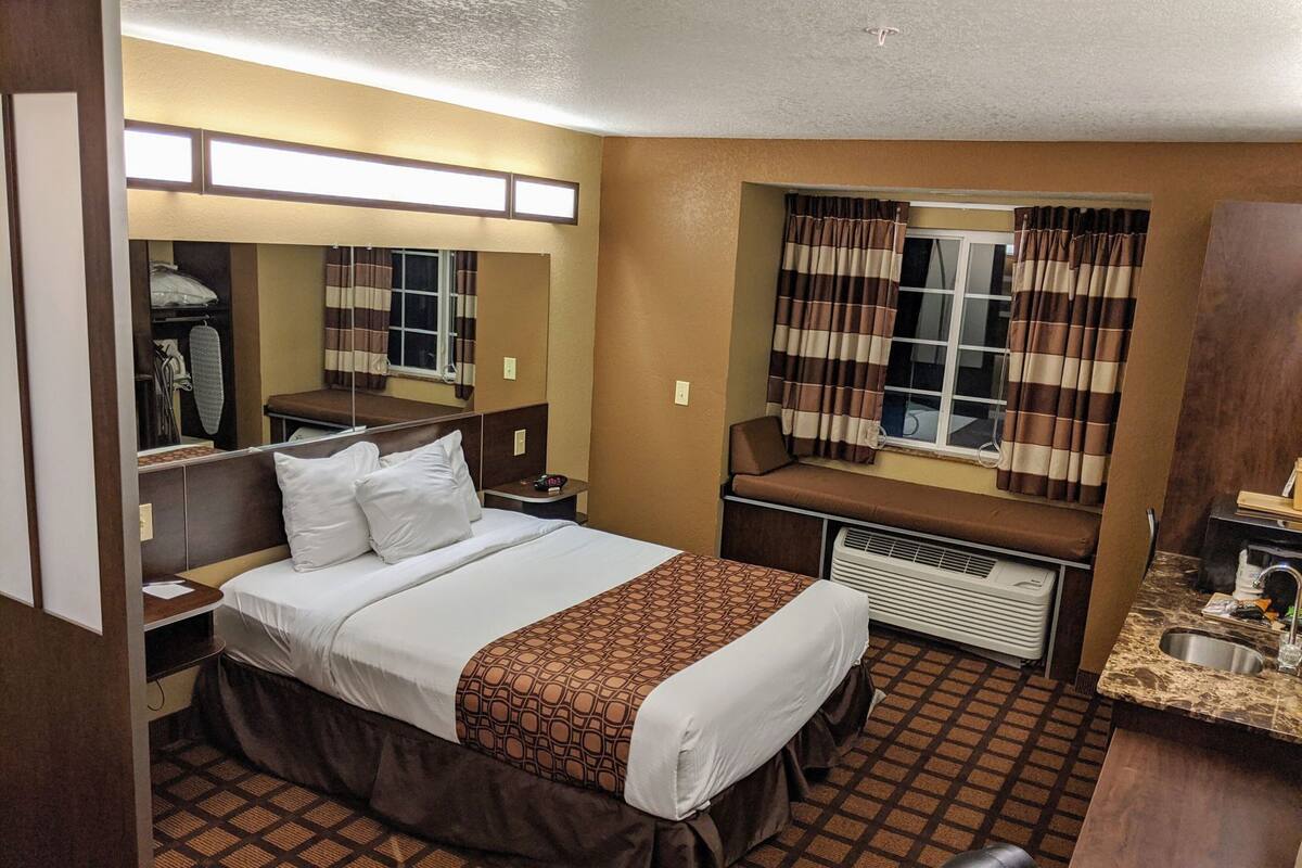 A hotel room with a bed, bedside table, mirror, window with curtains, and a small kitchenette area.