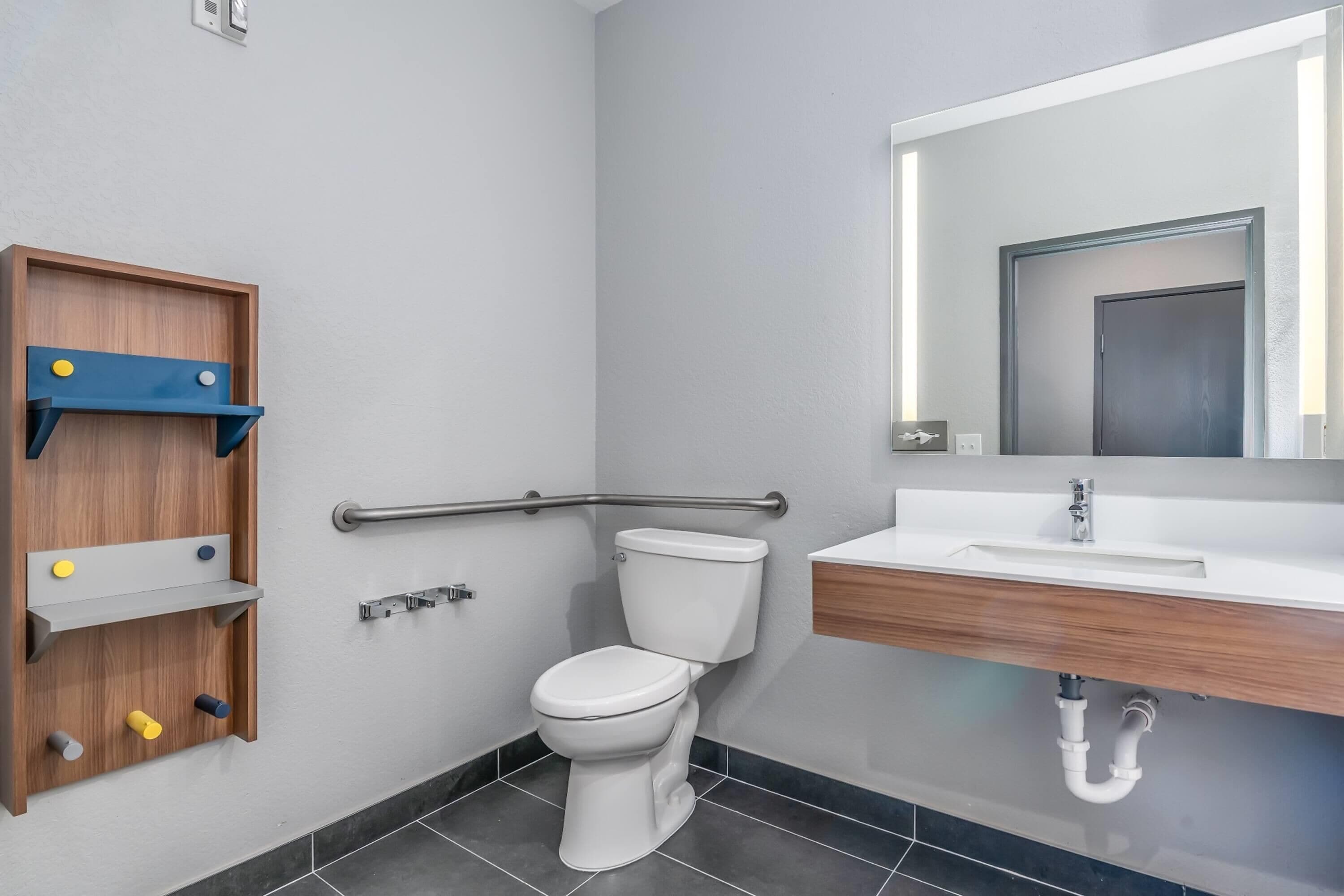 Une salle de bain moderne avec des toilettes blanches, un meuble-lavabo en bois doté d’un miroir et une barre d’appui pour faciliter l’accès.