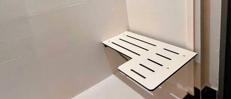 A corner of a room with white tiled walls and a black floor. There is a white, slatted shelf against the wall and a drain in the corner.