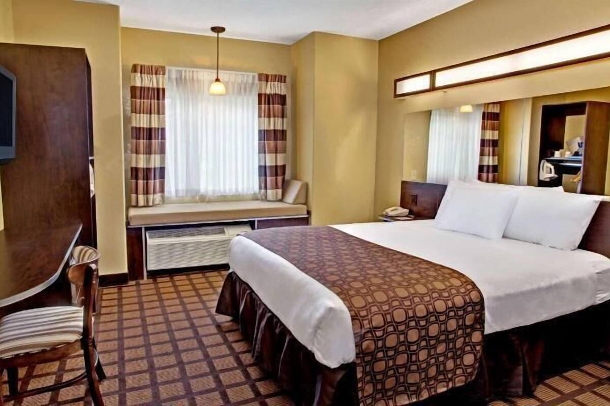 A hotel room with a bed, a desk, a chair, a TV, a window with curtains, and a mirror.