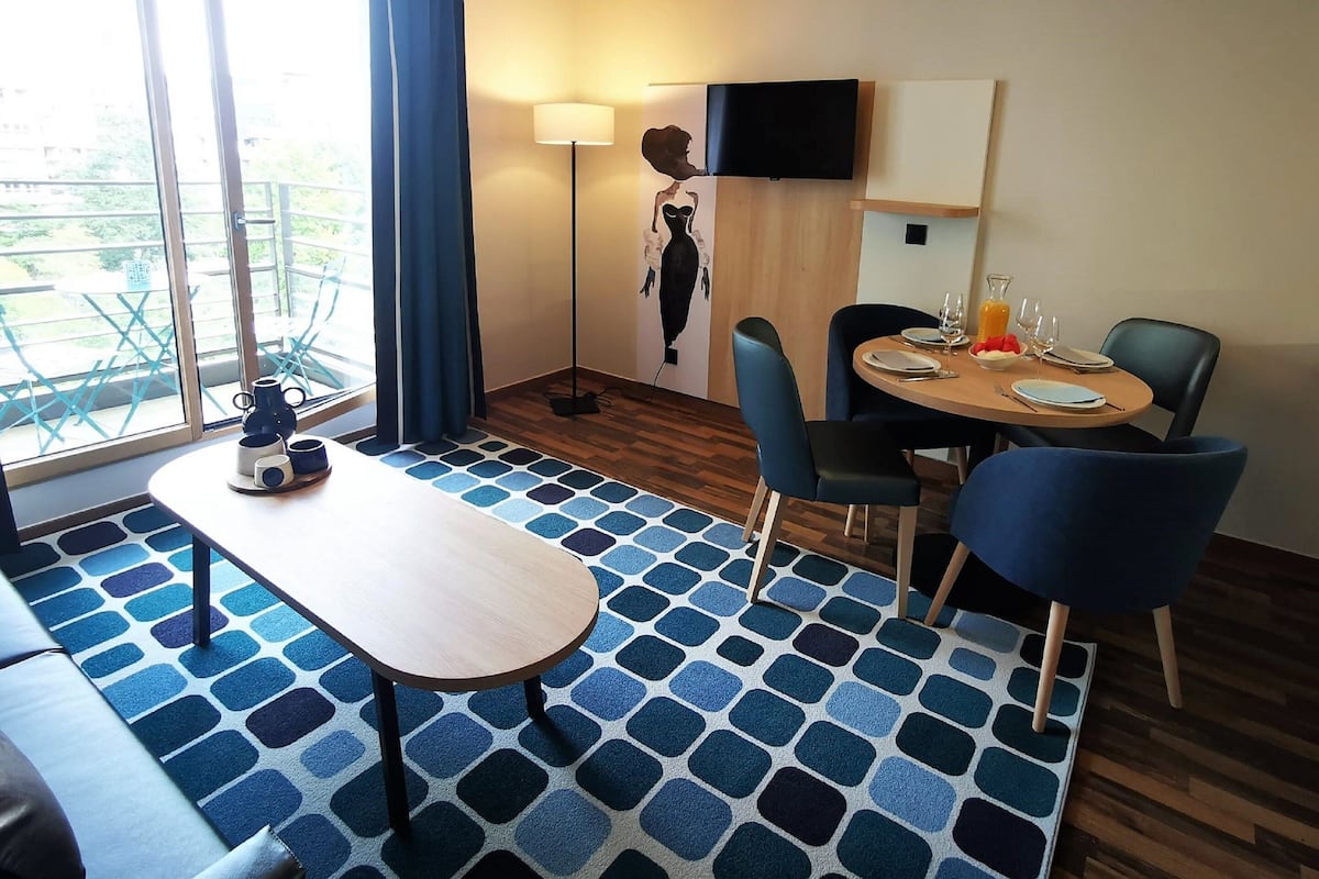 Hotelkamer met een eethoek, een bank, een televisie en een groot raam met gordijnen.