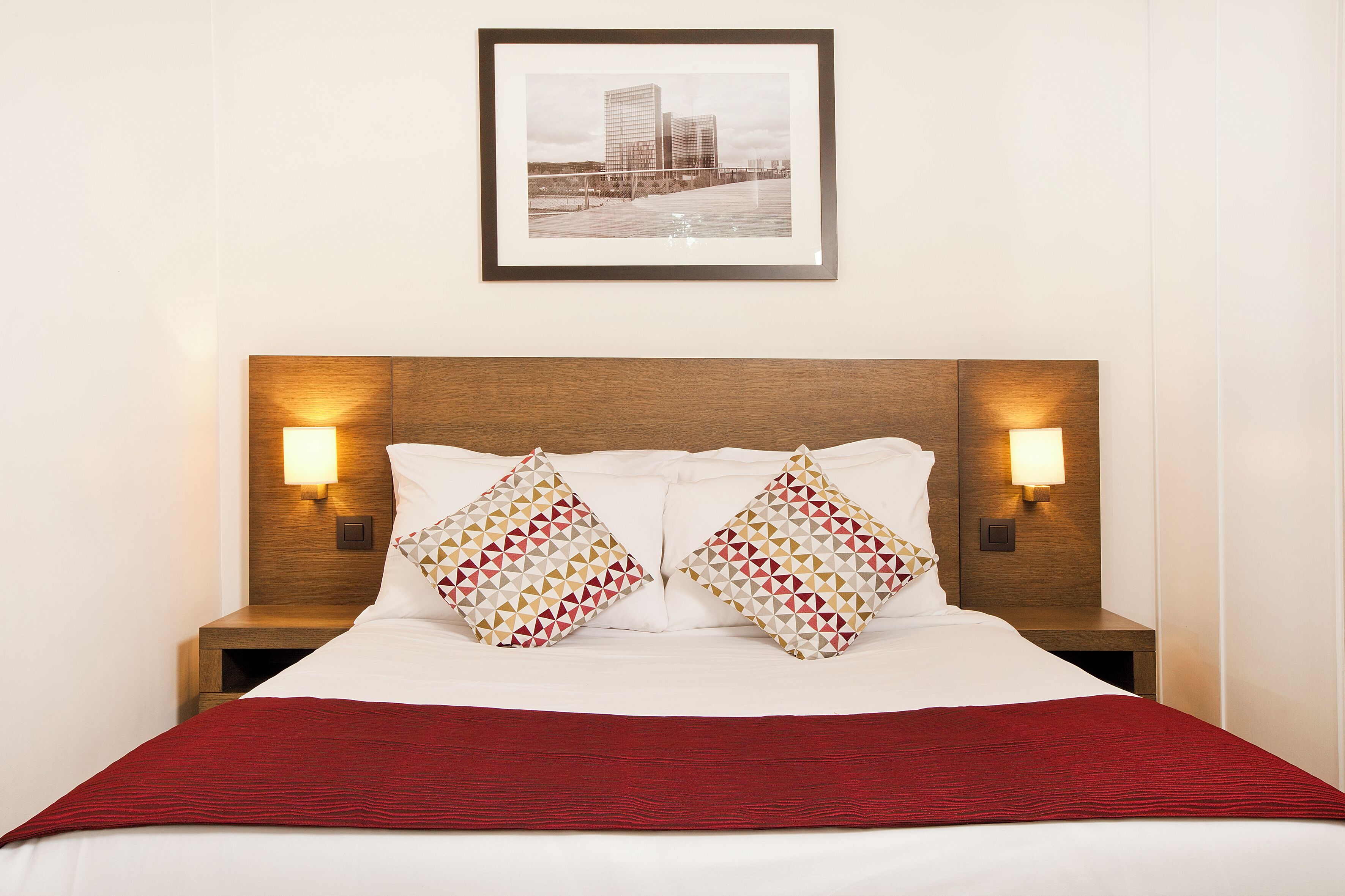 Una cama bien hecha con una colcha roja, dos almohadas estampadas y dos lámparas fijadas a la pared.